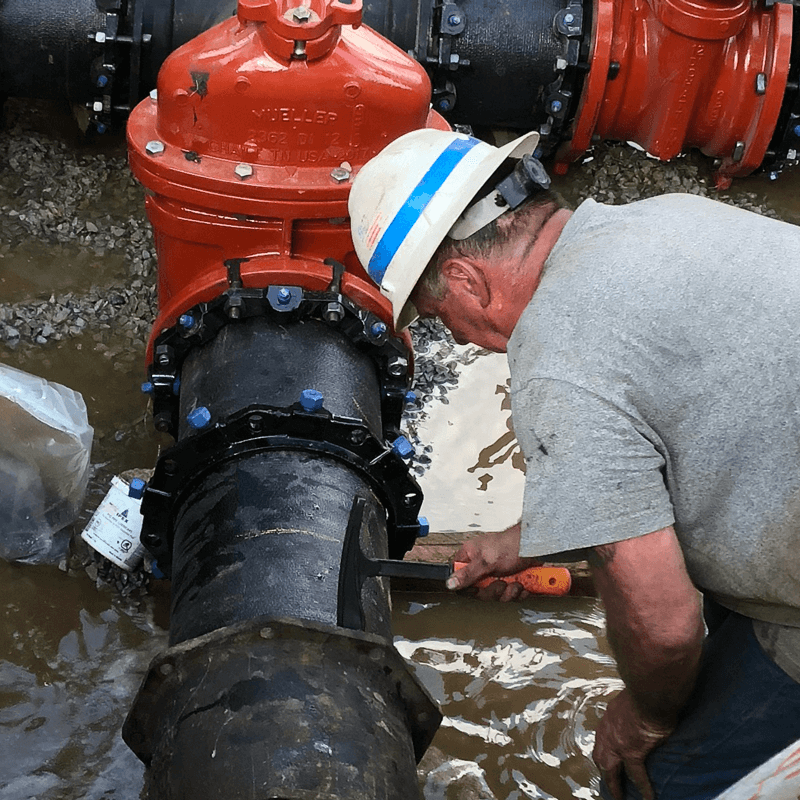 Man using a black hammer with an orange handle on a large pipe system outdoors in water